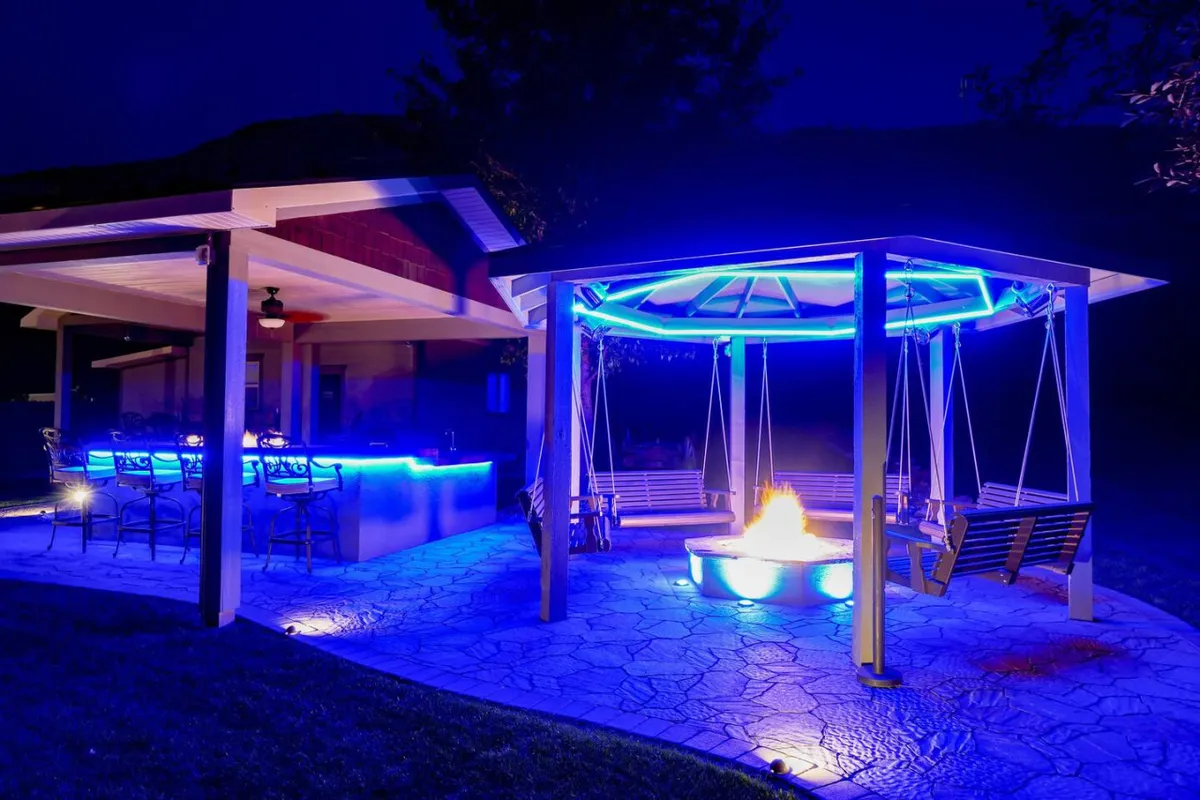 An illuminated gazebo stands welcoming under the night sky, surrounded by a cozy fire pit, bar area, and swing seating. The space is designed for evening gatherings, storytelling, and enjoying star-filled nights with loved ones.