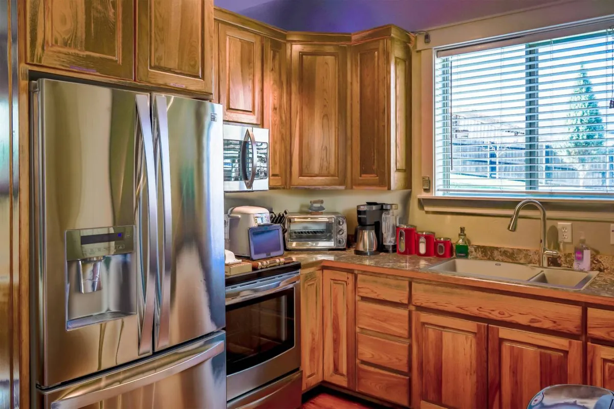 The image shows a bright, inviting kitchen with a sleek stainless steel refrigerator, sparkling granite countertops, a modern kitchen sink, and a large window with blinds that let in warm natural light. The space is perfect for cooking, baking, or gathering with loved ones.