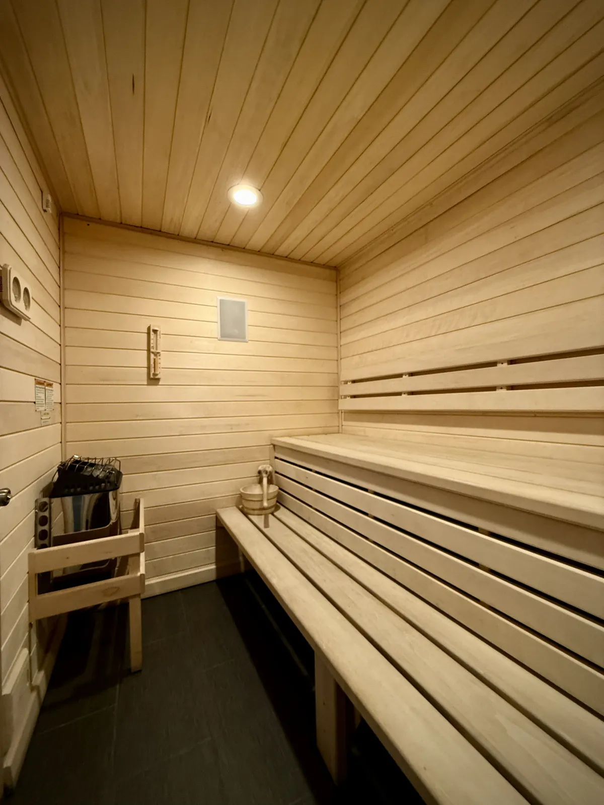 A cozy, warmly lit aspen cedar sauna interior with wooden benches, a traditional sauna stove, and spa essentials like a bucket and ladle, evoking a sense of wellness and intimate relaxation.