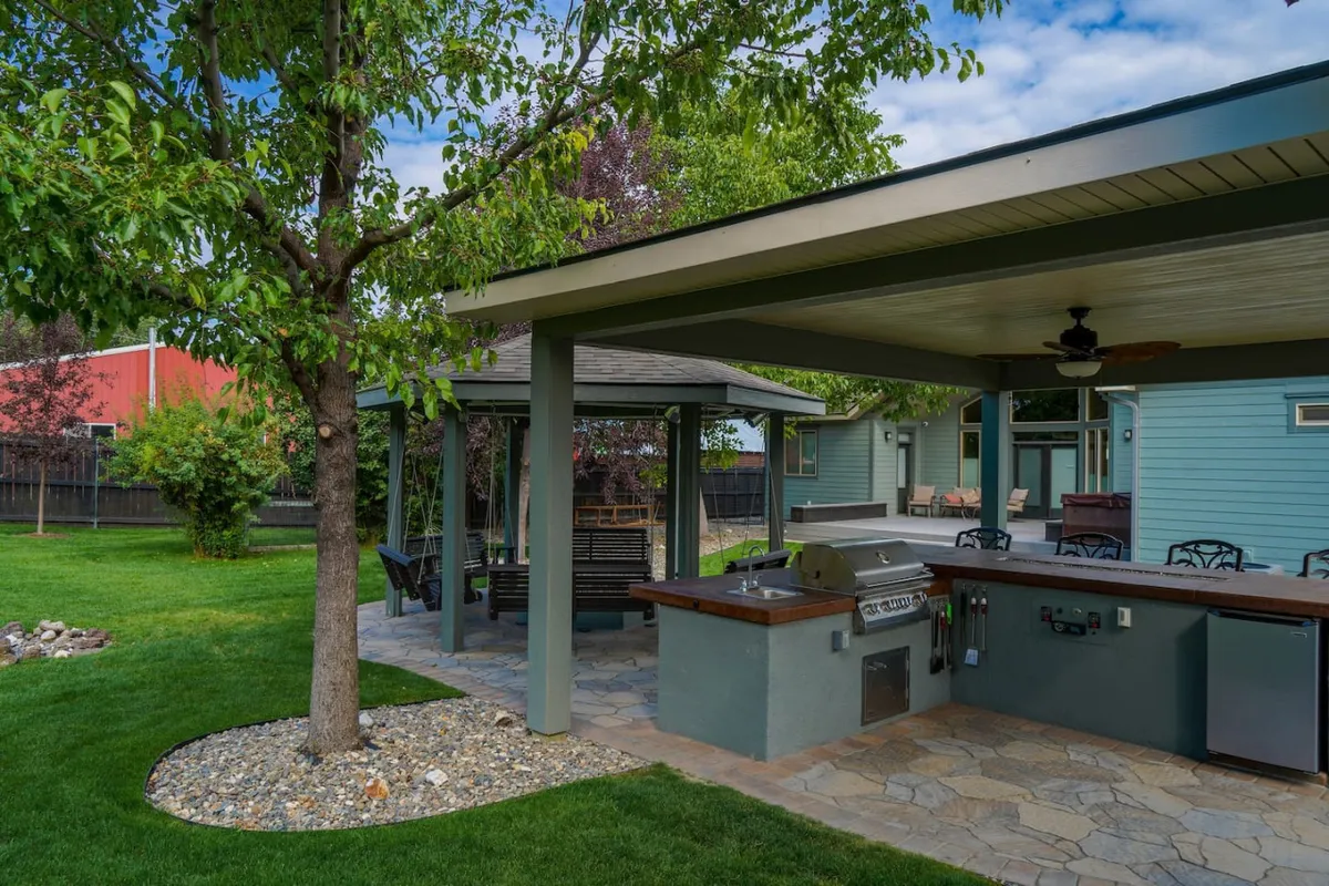 This shot features a well-equipped outdoor kitchen, a shaded patio area, and a nearby swing set, all surrounded by greenery. The scene feels inviting and upbeat, perfect for grilling, entertaining, and enjoying the outdoors.