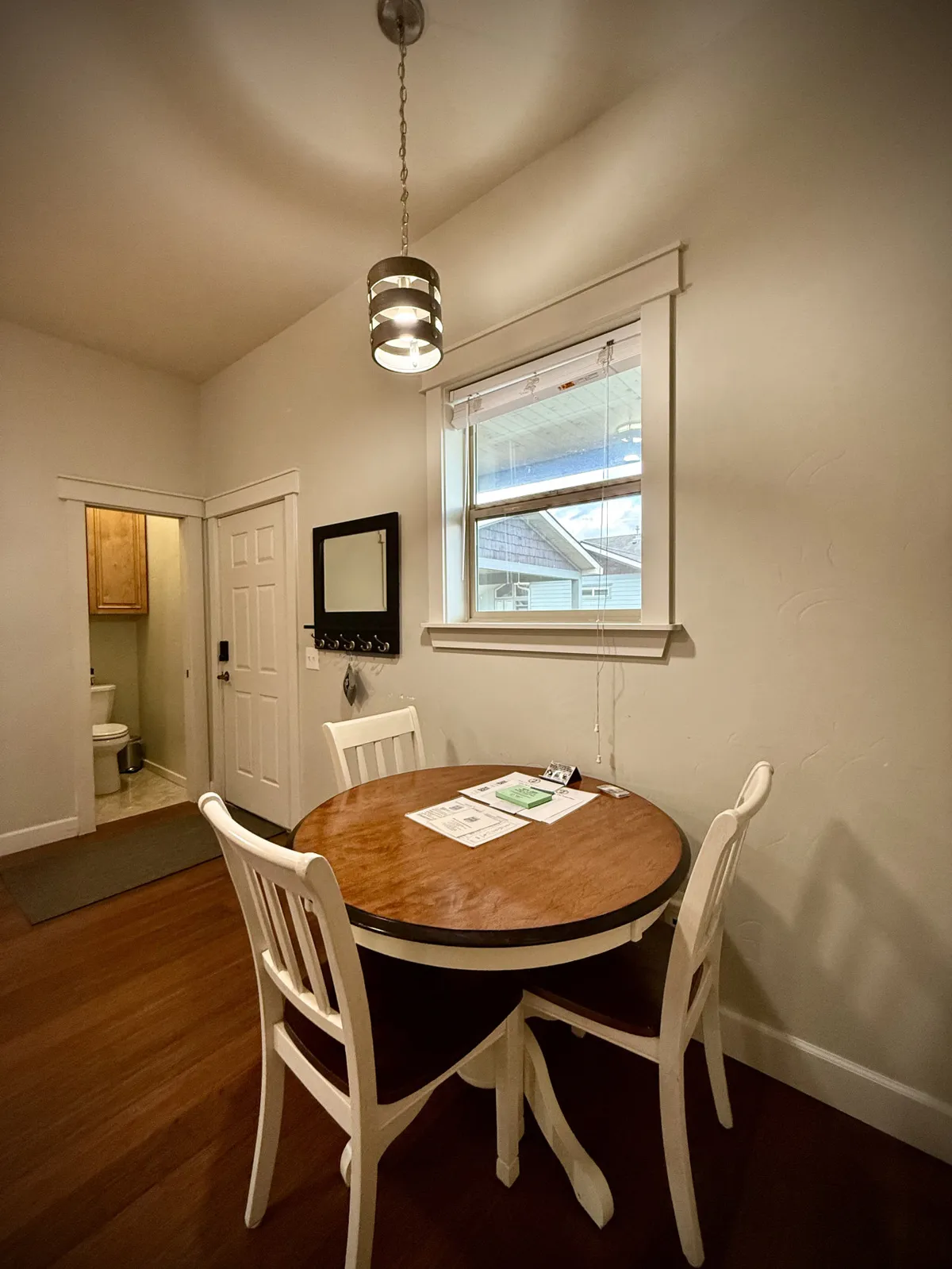 A cozy, round wooden dining table with white chairs, positioned beside a window dressed with blinds and a wall-mounted mirror reflecting the space. The nook feels welcoming, intimate, and perfect for sharing meals, coffee, or conversation, with natural light suggesting pleasant garden views.
