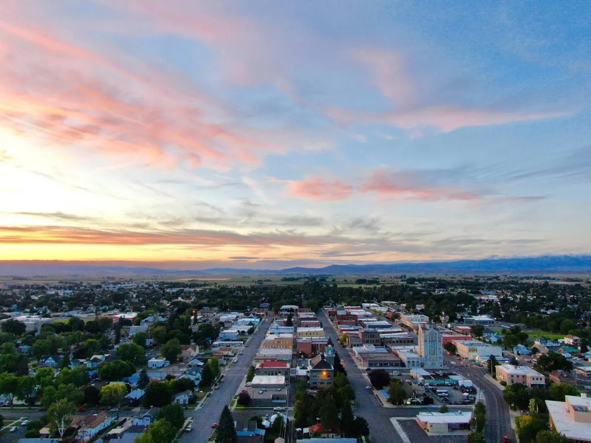 A wide, peaceful view over Baker City at sunset, with the town layout nestled between mountains in the distance. The image evokes a sense of tranquility and the small-town atmosphere, with hints of Main Street and its inviting shops and cafes.