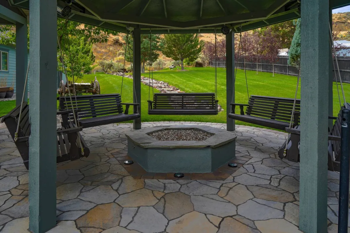A tranquil stone patio centers around a welcoming fire pit, with swing seats and a lush green lawn providing a cozy, communal setting. The layout invites quiet mornings with coffee as well as evening storytelling under the stars.