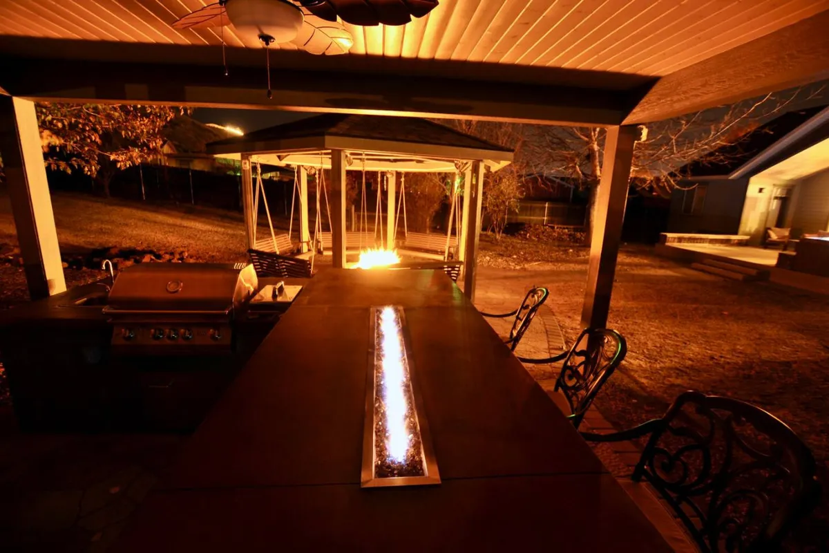 This image showcases an inviting outdoor kitchen grill and fire feature, with a swing set and a dining area visible. The scene is set for lively gatherings, with ample space for preparing meals, dining, and relaxing by the fire under the open sky.
