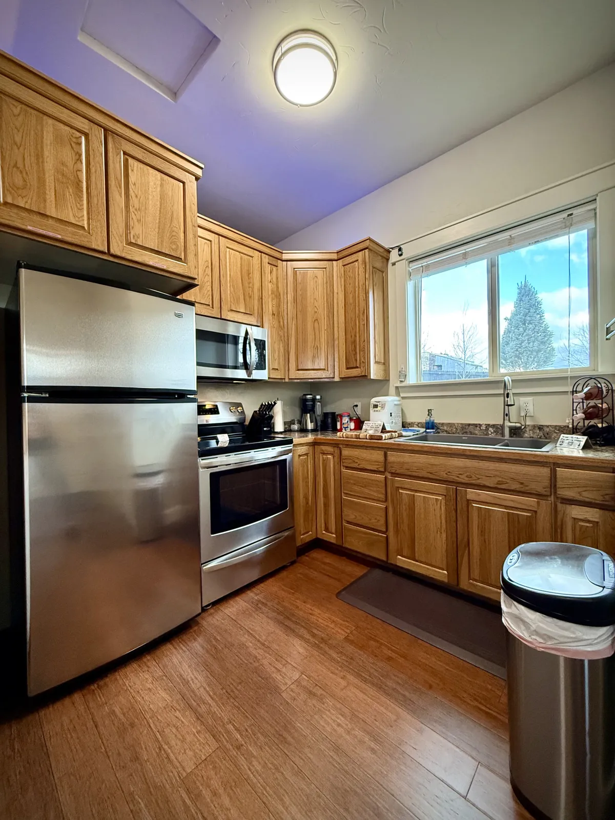 A warmly lit kitchen showcasing stainless steel appliances, wooden cabinetry, and a kitchen window that offers a view outside. The space radiates a homey atmosphere, suggesting ample room and tools for cooking and baking, as well as accommodating group meal prep or family gatherings.