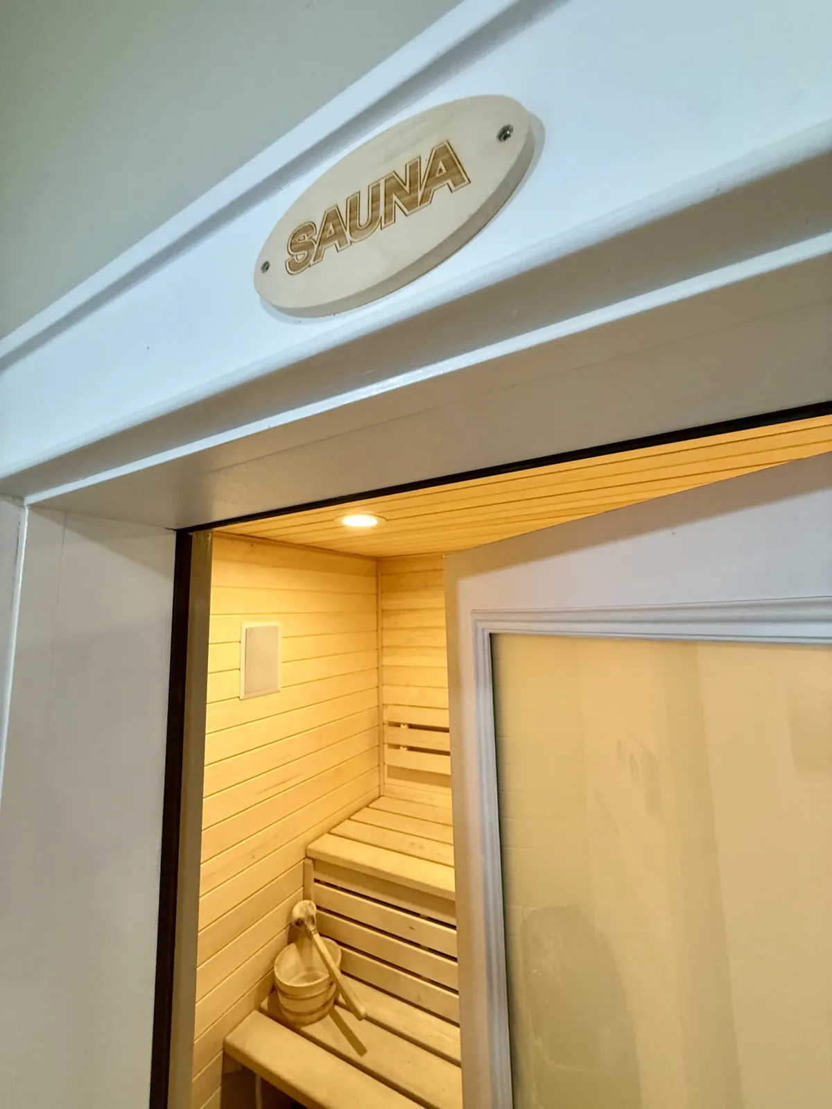 This image captures a tranquil cedar sauna featuring wooden benches, a wooden bucket, and a sauna sign. Warm wood tones and a cozy, intimate layout emphasize a spa-like ambiance ideal for relaxation and rejuvenation.
