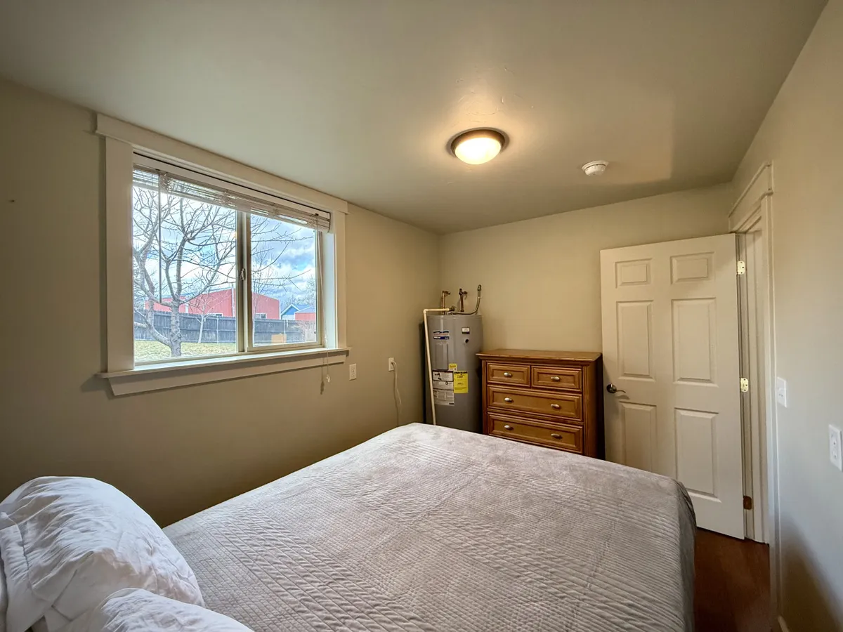 A serene bedroom setting with a comfortable bed, a window letting in natural light, a small dresser, and a refrigerator. The bedding and calm lighting evoke a tranquil and restful mood.